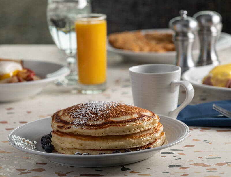 A close up of a stack of Georgie's pancakes served with a cup of coffee. Additional breakfast dishes and a glass of orange juice are in the background.