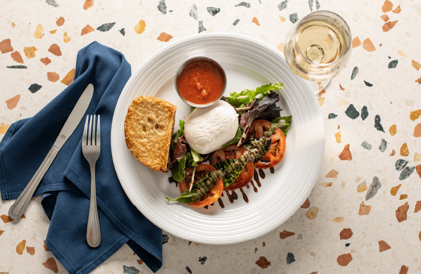 Georgie's Burrata Salad featuring Fresh Burrata cheese, sliced tomato, basil, pesto, and a balsamic reduction served with a side of marinara and fresh Parmesan garlic bread. The salad is paired with a glass of white wine.