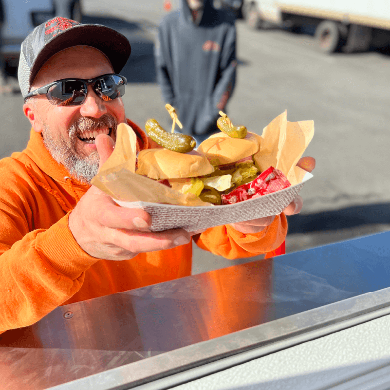 A smiling male customer taking his order from the counter of Georgie's Food Cart. The order consists of 2 sliders with pickles.